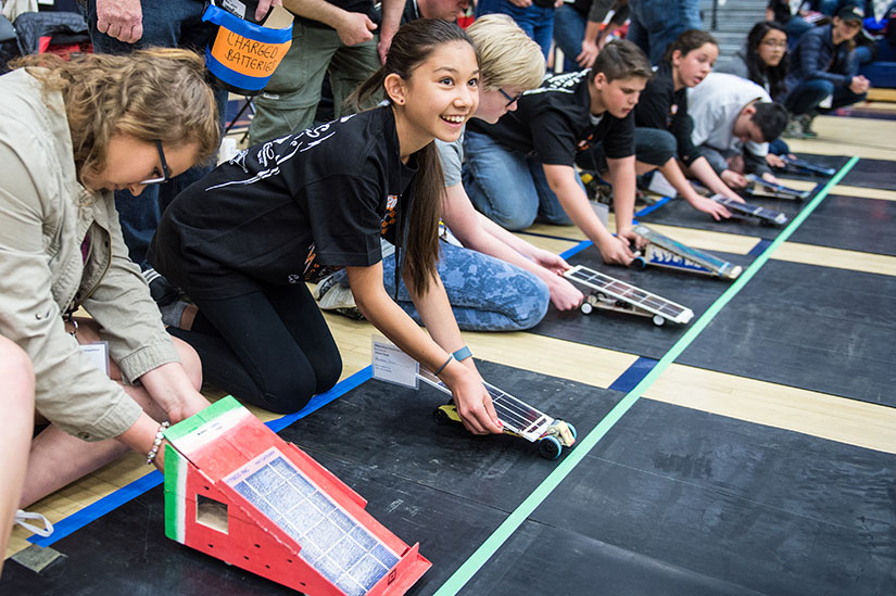 A row of students line up behind homemade electric toy cars. 