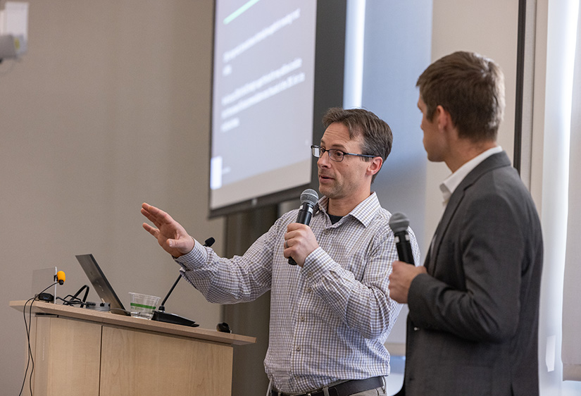 A man stands at a podium holding a microphone speaking with another man looking at him, also with a microphone, nearby.