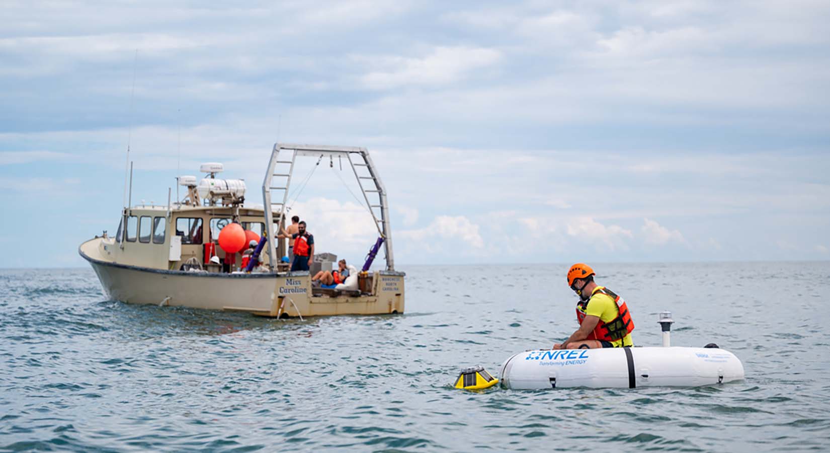 A man in an inflatable raft puts a device in the open water as a support ship watches nearby.