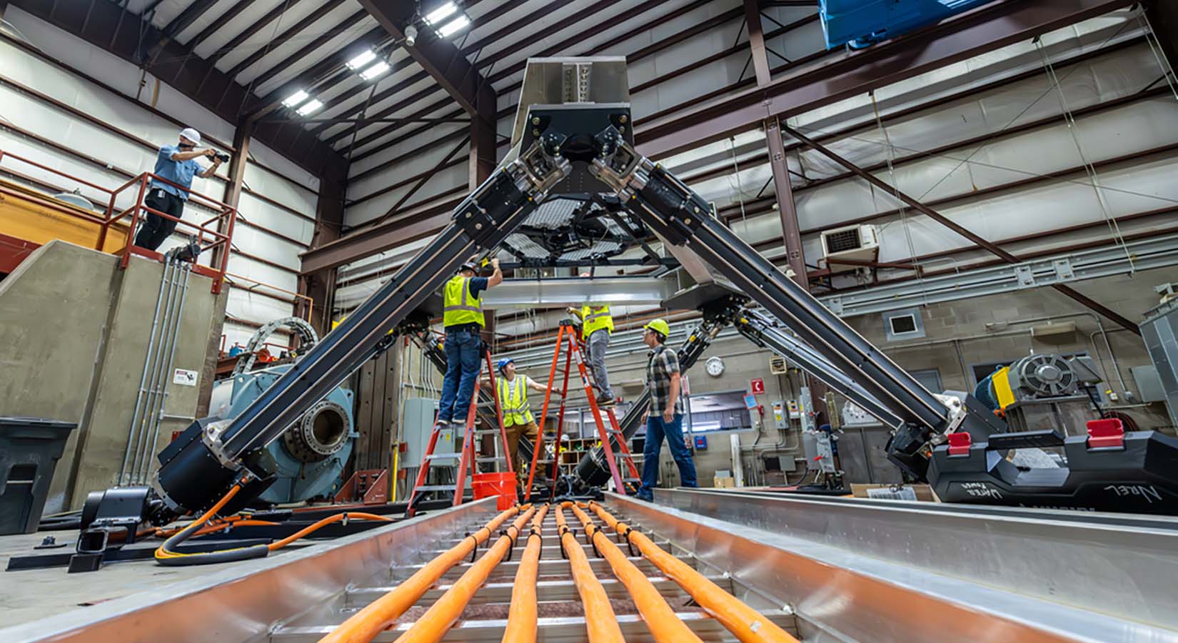 A track of wires leads underneath a platform held up by several large mechanical legs, while people in hard hats stand on ladders and examine the device.