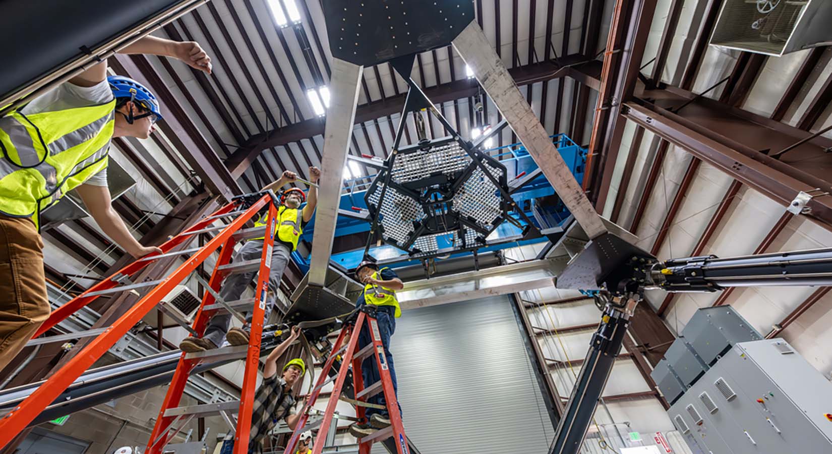 Looking up at a triangular platform that suspends a saucer-like device as people on ladders and high-vis vests examine the metal supports.