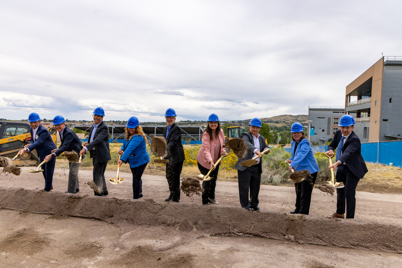 Nine people with shovels and hard hats toss dirt into a pile. 