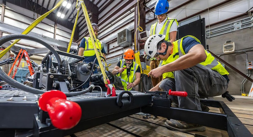 Four people in hard hats and safety vests work on a large metal piece of equipment