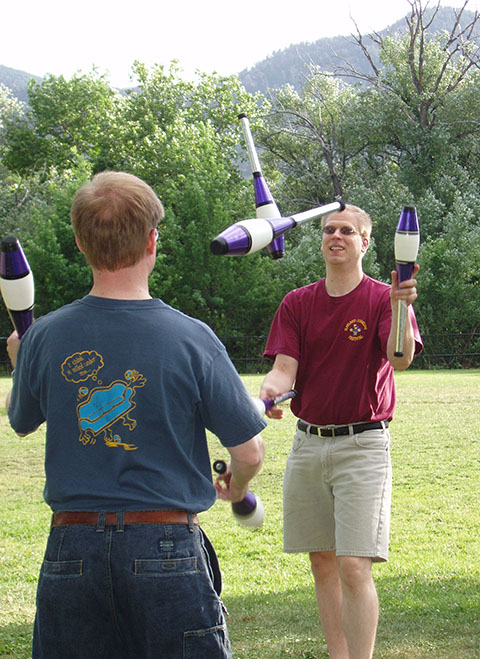 Two men juggle large juggling clubs outdoors.