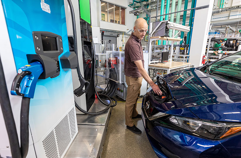 Man in safety glasses connects a charger to an electric vehicle inside a laboratory.