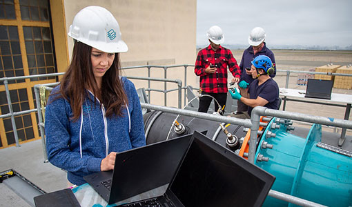 A person in a hard hat stands at a laptop in front of a big piece of hydropower machinery; three people in hard hats review something together