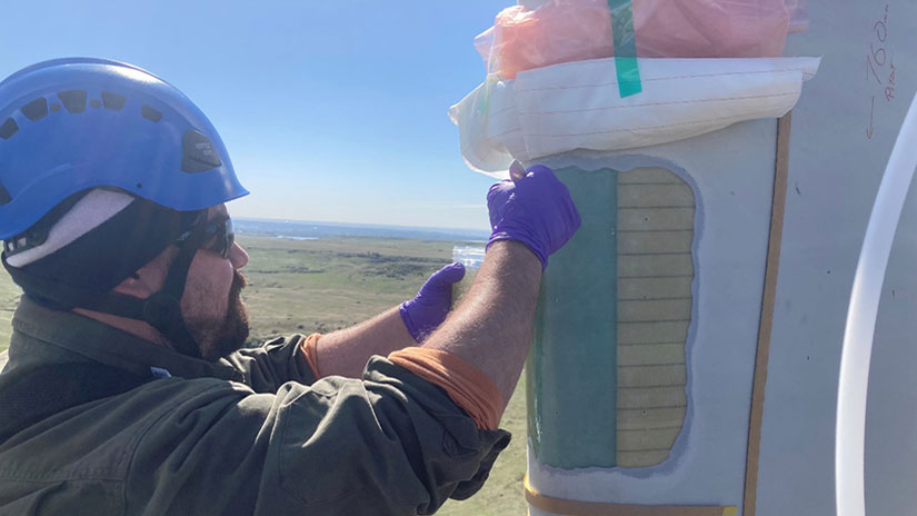 A person in a hard hat and safety gloves applying epoxy to the edge of a wind turbine blade