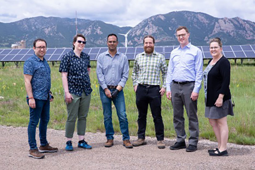 Six people standing outside in front of a row of solar panels and mountains