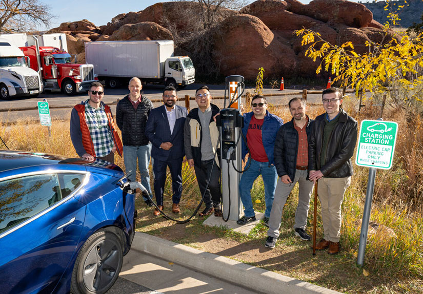 A group of people standing next to a charging electric vehicle. 