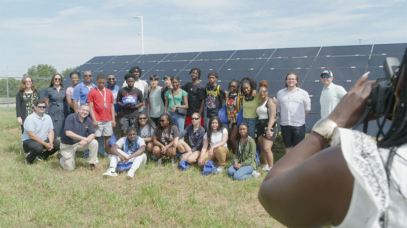 Group of people standing and sitting in front of solar panels