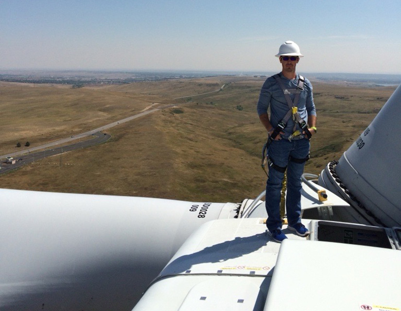 A photo of Heath Garrison in a helmet and harness on top of a wind turbine at NREL’s Flatirons Campus. 