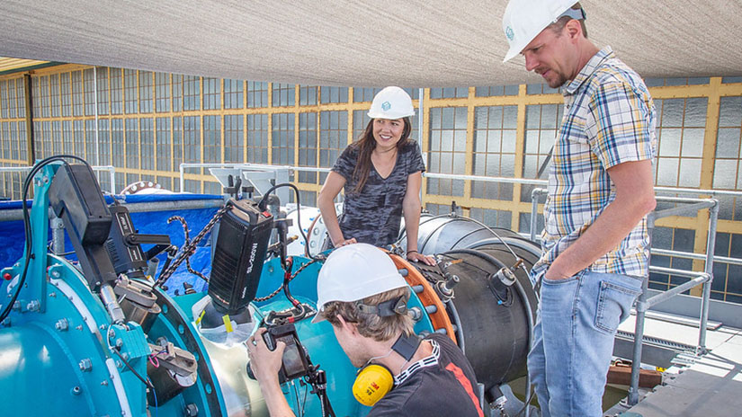 Three people in hard hats work around a large metal pipe.