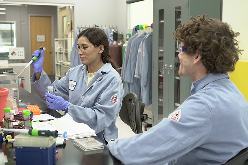 Two people in blue lab coats in a lab. One of them, a woman, is collecting material to put into a plastic tube.