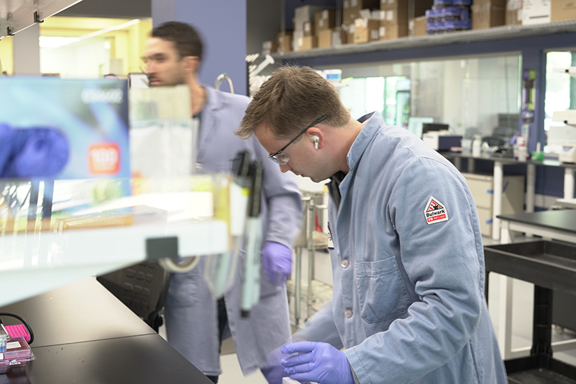 A man in a blue lab coat in a laboratory looks down at something as another man walks in the background.