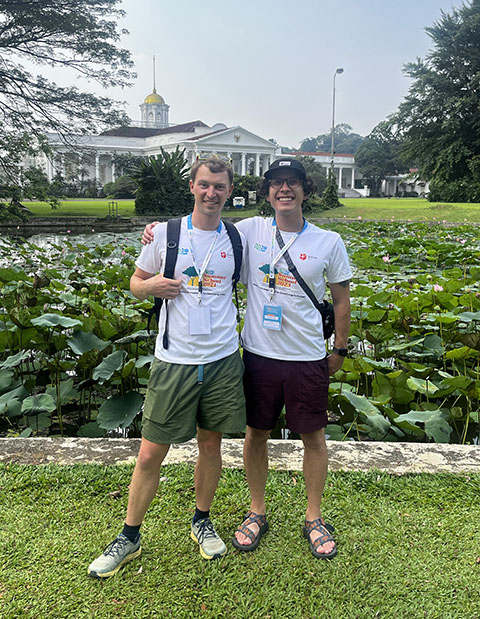 Two people stand in front of a garden with a palace in the background.