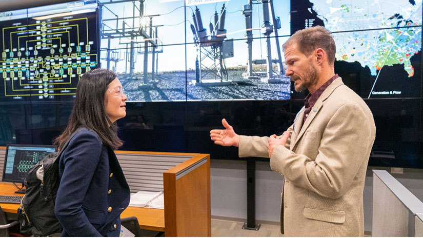 Two people talking in front of screens showing an electrical substation and visualizations of the U.S. power grid