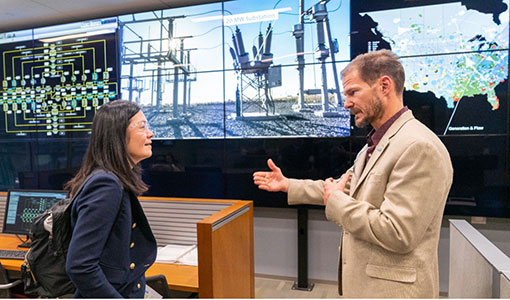 Two people talking in front of screens showing an electrical substation and visualizations of the U.S. power grid