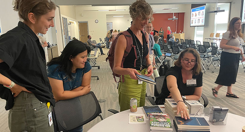 A person sits at a table with books as others  stand around the table.