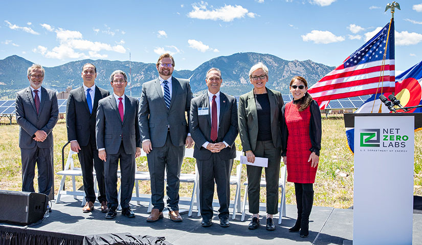 Seven people pose on an outdoor stage next to a flag. 