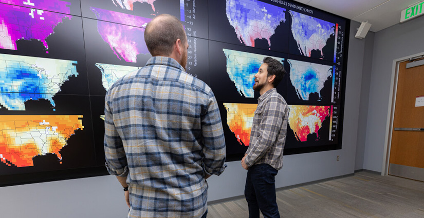 Two people stand and chat in front of screens showing different-colored data layers over maps of the United States.