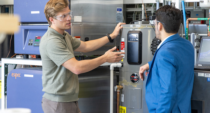 Two people stand in front of piece of HVAC equipment.