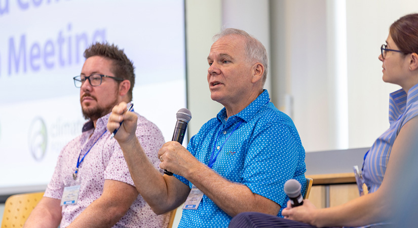 Three people sit in the front of a room with microphones, one in the process of speaking.