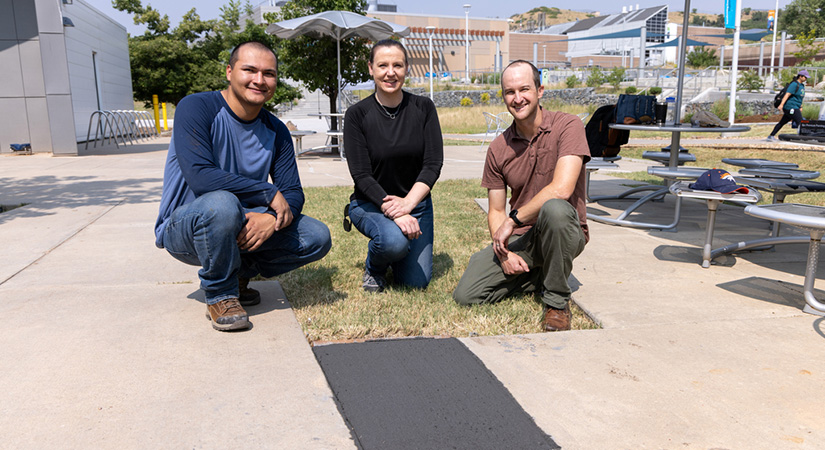 Three people crouch behind a slab of concrete that is a darker gray color than the surrounding concrete.