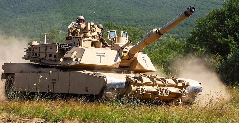 Several U.S. soldiers drive an M1 Abrams tank across a green field, kicking up a cloud of dust.