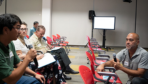 Two people hold microphones in a room with chairs and projector screen