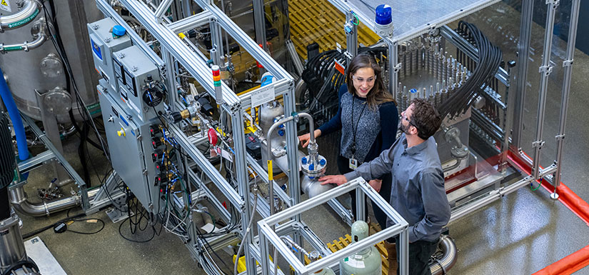 Two researchers talk to each other while working in a laboratory with large equipment.