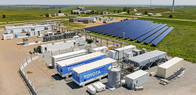 An aerial view of NREL’s Flatirons campus, with large hydrogen equipment in the foreground, a solar panel field in the middle ground, and grass and a wind turbine in the background.