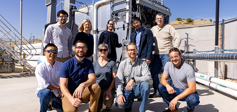 Ten researchers smile in front of a large piece of hydrogen fueling equipment outdoors.