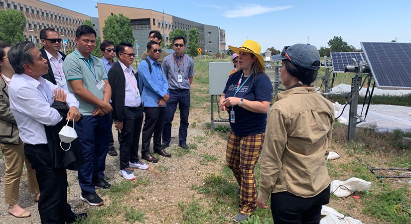 A group of individuals stand outside, near a solar PV installation. Two women in hats are presenting at the front.