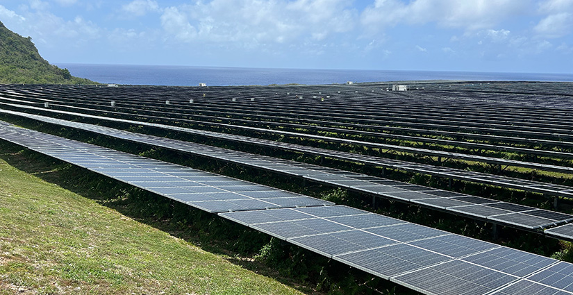 Solar panels in a grass field with ocean in the distance.