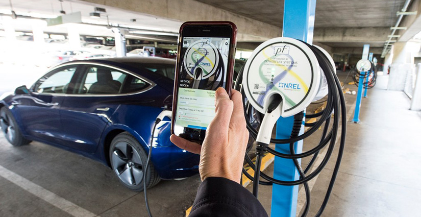 A person holds a smartphone showing a smart charge management app in front of a plugged-in blue electric car.