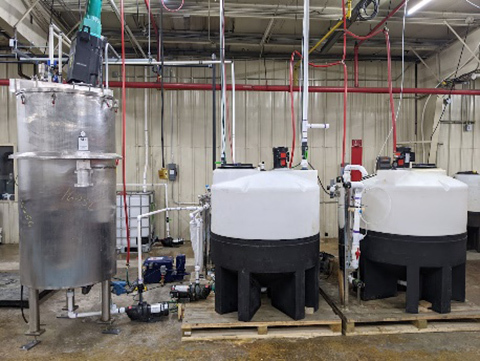 A tall metal tank stands next to two round plastic tanks in a warehouse. Multiple pipes and tubes run between the tanks to facilitate rare earth purification.