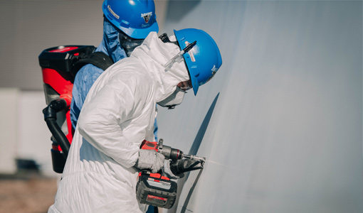 A student wearing protective gear drills into a wind turbine blade to cut out a square sample while another person stands behind them to oversee the work.