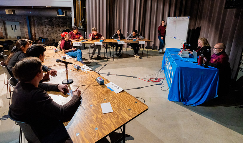 On a stage, two tables of students face a table with two judges. 