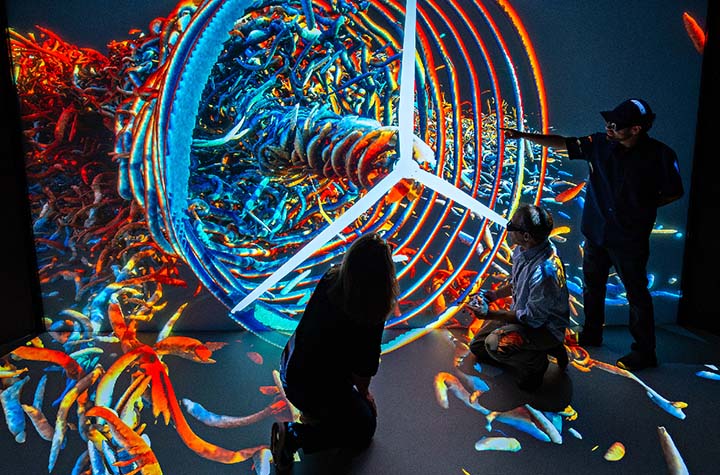 three researchers in silhouette interacting with a multicolor 3D visualization featuring a wind turbine