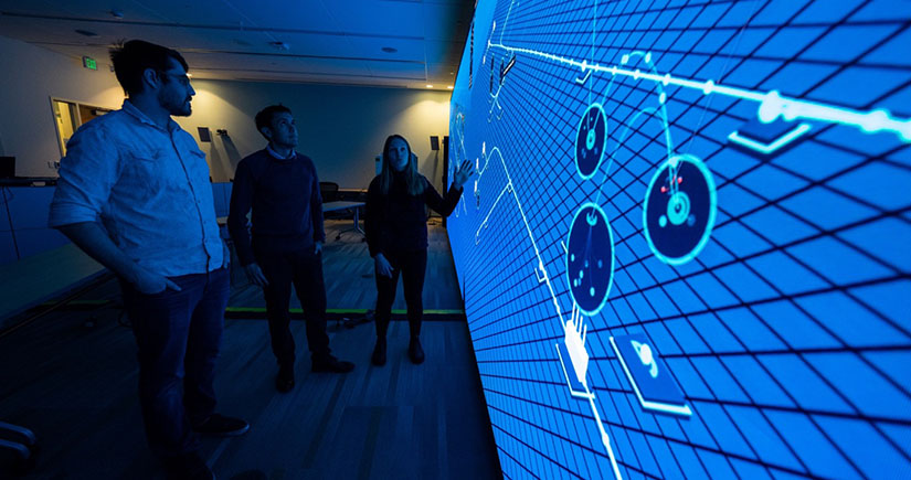 Three people stand in a dark room looking at a large wall display computer screen.