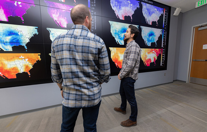 Two people stand and chat in front of screens showing different-colored data layers over maps of the United States.