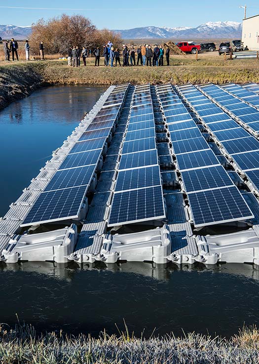 An array of connected solar panels seen in profile floating in a pond with mountains in the background
