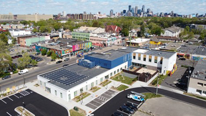 A neighborhood in the Twin Cities area of Monnesota. In the foreground, a one-story small business building with a rooftop solar installation. In the background, a downtown skyline.