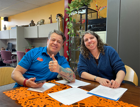 Two smiling people sitting at a table in an office. One is giving a thumbs-up.