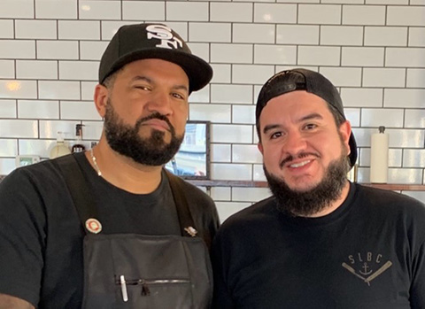 Two people stand side by side in front of a tiled wall. Both have beards and hats.