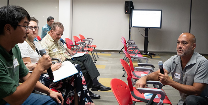 A person who is sitting in a row of chairs speaks into a microphone, while another person kneeling in the row in front of him listens.