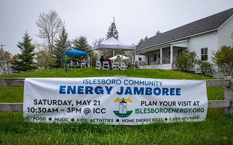 A banner on a fence that reads Islesboro Community Energy Jamboree with tents, chairs, and a house in the background.