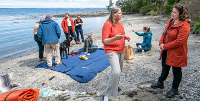 Eight people interacting on a beach with a blanket on the ground and trees in the background.