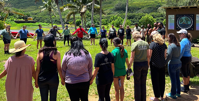 About 25 people stand in a circle holding hands beneath palm trees.