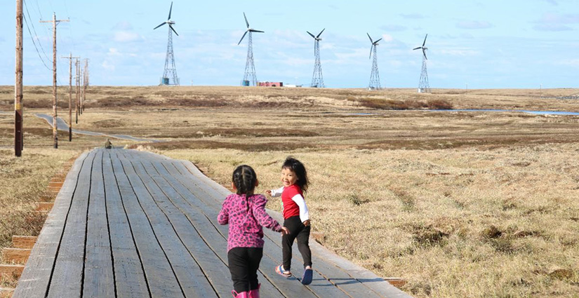 Two children run down a boardwalk in an open field with wind turbines and power lines in the background.